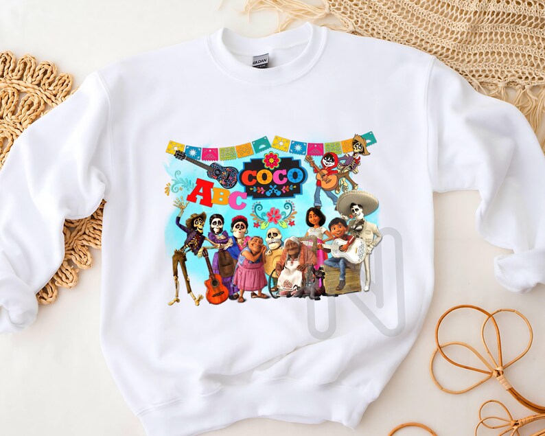 May include: White sweatshirt with a colorful graphic print of characters from the movie Coco. The print includes the characters Miguel, Hector, Dante, and others. The text "ABC COCO" is also included in the print.