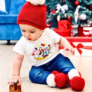 May include: A toddler wearing a red knit hat with a white pom pom, a white onesie with a colorful Disney character print, and blue jeans. The toddler is holding a pink toy and sitting on a wooden floor.