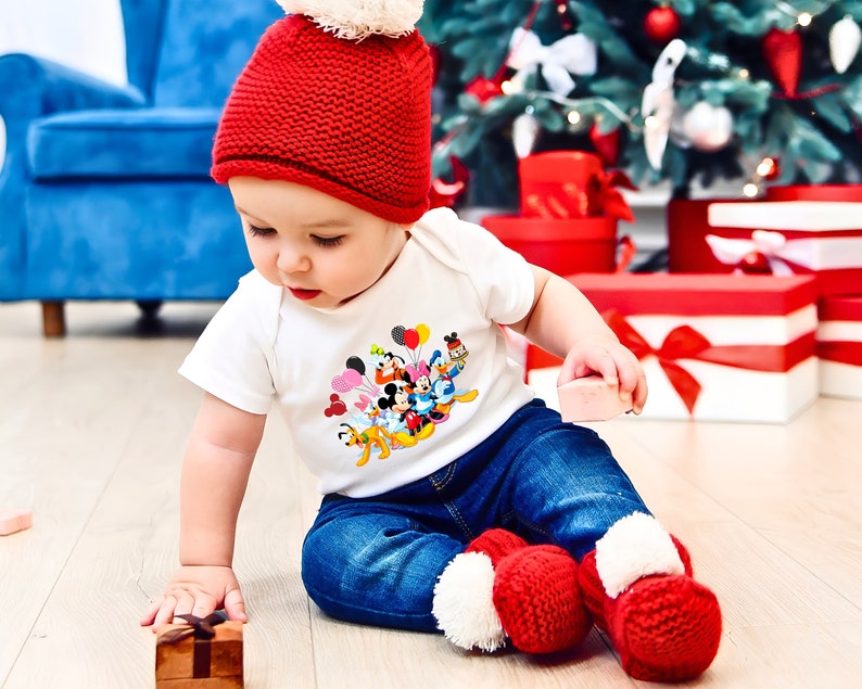 May include: A toddler wearing a red knit hat with a white pom pom, a white onesie with a colorful Disney character print, and blue jeans. The toddler is holding a pink toy and sitting on a wooden floor.
