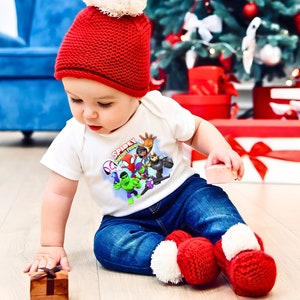 May include: A baby wearing a white onesie with a red Santa hat and blue jeans. The onesie has a graphic of Spiderman and his friends with the text "Spidey with Friends".