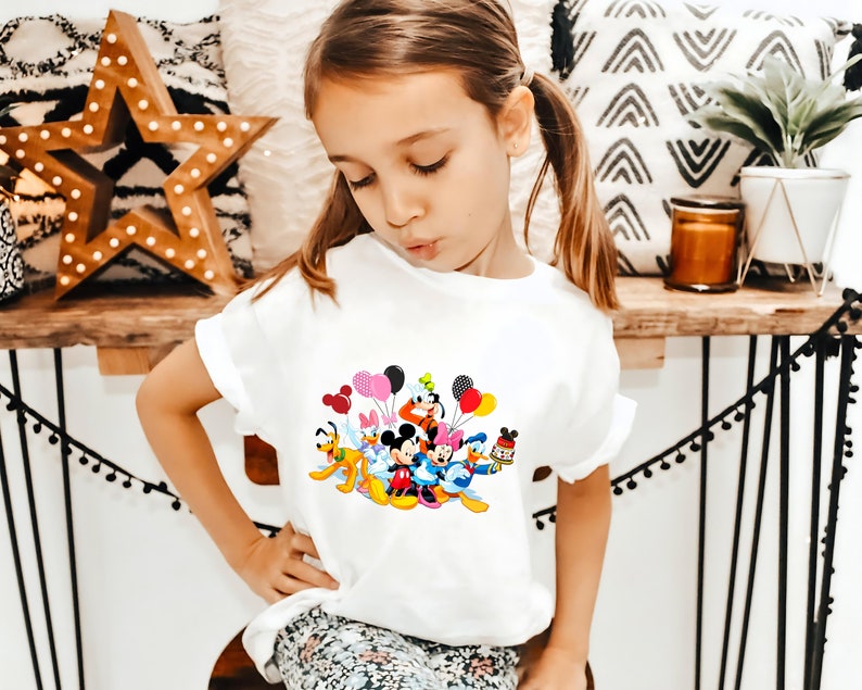 May include: A white t-shirt with a colorful graphic of Mickey Mouse and friends holding balloons. The graphic features Mickey Mouse, Minnie Mouse, Donald Duck, Goofy, Pluto, and other Disney characters.