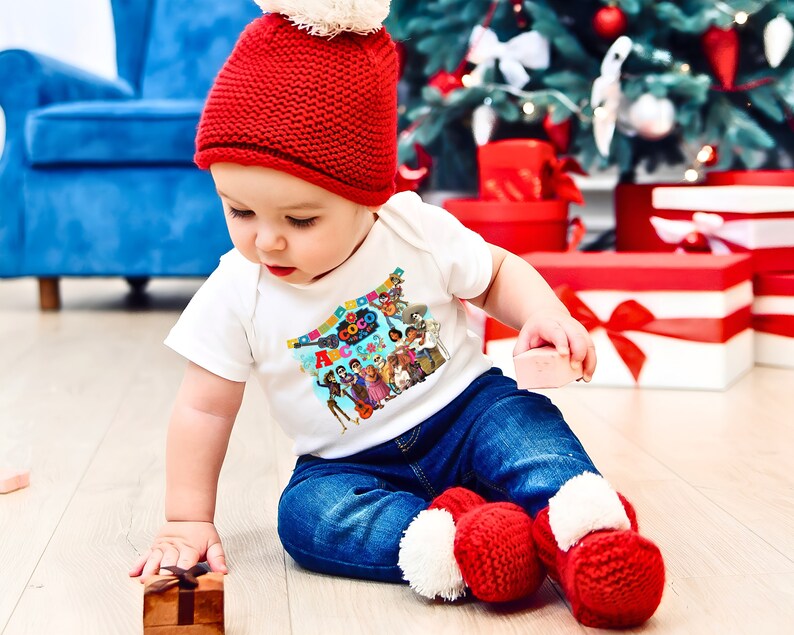 May include: A baby wearing a white onesie with a colorful Coco movie graphic, red knit hat, blue jeans, and red and white knit booties. The baby is sitting on a wooden floor in front of a Christmas tree.