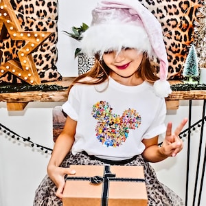 May include: A young girl wearing a white t-shirt with a colorful Mickey Mouse design, a pink Santa hat, and a leopard print skirt. She is holding a wrapped gift box with a black ribbon.