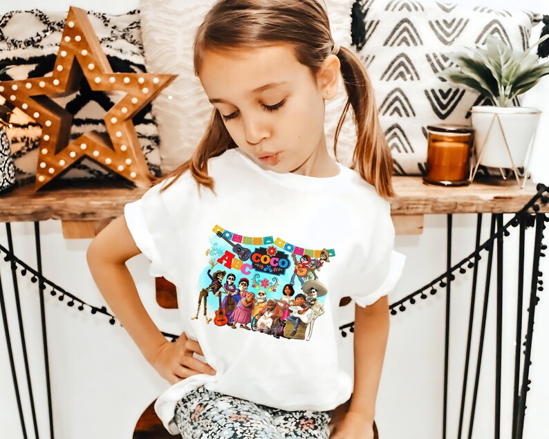 May include: White t-shirt with a colorful graphic of characters from the movie "Coco" and the text "ABC Coco The Day of the Dead".
