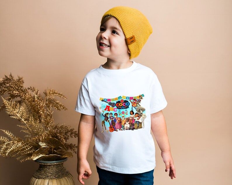 May include: A white t-shirt with a colorful graphic of characters from the movie "Coco" and the text "Coco" and "ABCs of Life". The shirt is being worn by a child wearing a yellow beanie.