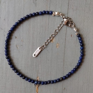 Könnte beinhalten: Ein zartes Armband mit kleinen, dunkelblauen Perlen, die aufgefädelt sind. Das Armband hat eine silberfarbene Kette und einen Verschluss mit verstellbarer Länge. Die Perlen sind gleichmäßig angeordnet und bilden ein schlichtes, elegantes Design. Das Armband ist etwa 18 cm lang.