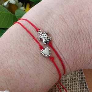 Può includere: Due braccialetti di corda rossa con ciondoli d'argento. Un ciondolo è una tartaruga e l'altro è un cuore.