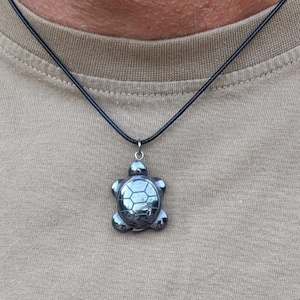 May include: A turtle pendant necklace. The pendant is a dark, metallic grey, turtle-shaped charm with a detailed shell design. The necklace has a black cord and a silver-coloured clasp. The background is a tan-coloured shirt.