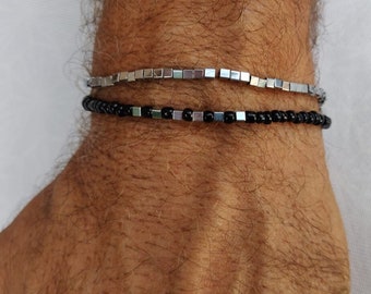 Puede incluir: Dos pulseras en una muñeca. La pulsera superior es plateada con cuentas cuadradas. La pulsera inferior es negra con cuentas redondas.