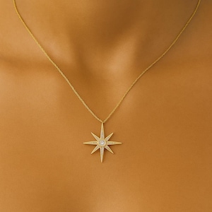 Può includere: Una collana color oro con un ciondolo a forma di stella. Il ciondolo presenta un design circolare centrale e molteplici raggi appuntiti, ciascuno ornato da piccoli accenti scintillanti. La collana è mostrata su uno sfondo color pelle.