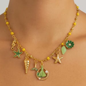 Può includere: Collana dorata con perline gialle e ciondoli a tema marino. I ciondoli includono un cavalluccio marino, una stella marina, una conchiglia, una coda di sirena, un nappa, un fiore e una foglia. I ciondoli sono verdi, dorati e bianchi.