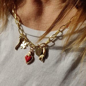 Può includere: Collana a catena dorata con più ciondoli, tra cui un cuore rosso, un quadrifoglio, un peperoncino, una mano con il gesto 'rock on' e un ciondolo circolare con la lettera 'R'.