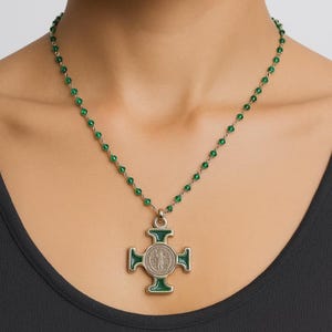 Può includere: Una collana color argento con un ciondolo a forma di croce verde e una catena di perline verdi abbinata. Il ciondolo a croce ha un centro color argento con un design dettagliato e dettagli in smalto verde. La collana è indossata intorno al collo.