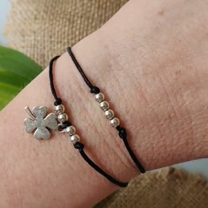 Può includere: Braccialetto con cordino nero con perline color argento e un ciondolo a forma di quadrifoglio. Il braccialetto è indossato su un polso, mostrando il contrasto tra il cordino scuro e gli accenti metallici lucenti. Un gioiello semplice ed elegante.
