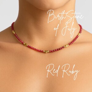 Può includere: Una collana di rubini rossi con dettagli dorati e un ciondolo a forma di cuore. La collana è composta da piccole perline rosse sfaccettate e perline color oro. Il testo sull'immagine recita "Birth Stone of July" e "Red Ruby".