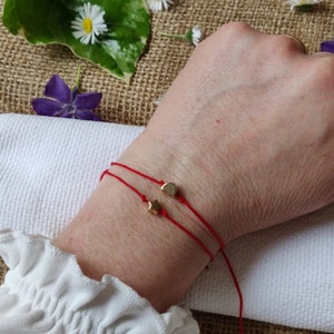 Puede incluir: Dos pulseras de hilo rojo con abalorios dorados, una en forma de corazón y otra en forma de estrella, se llevan en la muñeca. El fondo incluye flores y una superficie texturizada. Las pulseras son delicadas y sencillas.