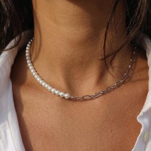 Può includere: Una collana dal design diviso in due parti. Una metà è composta da una fila di perle bianche lisce e rotonde, mentre l'altra metà è formata da maglie metalliche color argento con una forma rettangolare allungata.