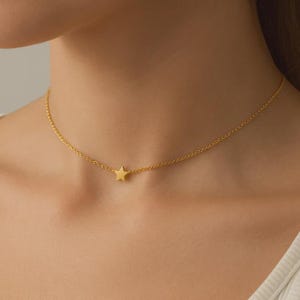 May include: A delicate gold-colored necklace featuring a small star pendant. The chain is fine and the star is centered, creating a minimalist and elegant design. The necklace is worn around the neck.