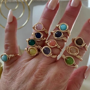 Può includere: Una collezione di anelli avvolti in filo d'oro, ognuno con una gemma colorata diversa. Le gemme includono rosa, blu, verde, giallo, arancione, viola e marrone. Gli anelli sono esposti su una mano con unghie lunghe e lucide.