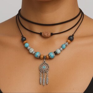 Può includere: Un set di collane a strati con un choker nero e una collana di perline. La collana di perline ha perline turchesi, argentate e di legno, con un ciondolo acchiappasogni e piume pendenti. Il choker ha una perla di legno.