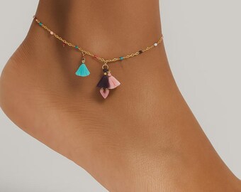 May include: Gold-coloured anklet featuring colourful beaded accents and three tassel charms. The charms include a turquoise tassel, a dark purple tassel, and a pink tassel with a heart-shaped charm. The anklet is worn on a foot.