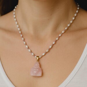 May include: A delicate necklace featuring a string of white pearls and small pink beads. A rose quartz Buddha pendant hangs from a gold-colored clasp. The necklace is worn around the neck.