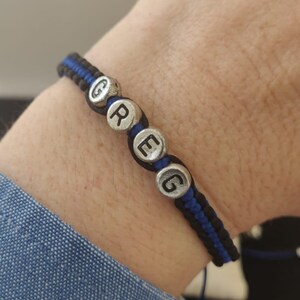 Può includere: Un braccialetto intrecciato nero e blu con il nome "GREG" scritto con perline argentate. Il braccialetto è su un polso. Lo sfondo è di colore chiaro.