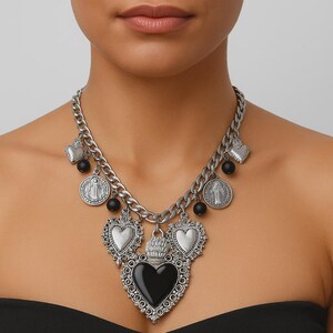 Può includere: Collana color argento con una catena spessa e vari ciondoli. I ciondoli includono ciondoli a forma di cuore, medaglioni rotondi e perline nere. Il ciondolo centrale è un grande cuore ornato con un centro nero.