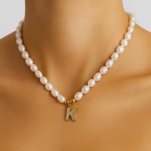 Può includere: Una collana di perle bianche con un ciondolo dorato a forma di lettera "K". La collana è composta da perle ovali e ha una chiusura dorata. Il ciondolo è incastonato con piccole gemme trasparenti.