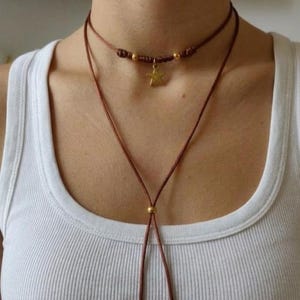 May include: A layered necklace with a brown cord. The top layer is a choker with gold beads and a gold star pendant. The second layer is a longer cord with a gold bead at the bottom. The person is wearing a white vest top.