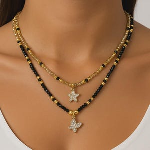 Può includere: Collana a due fili con perline dorate e nere. Il filo superiore ha un ciondolo a forma di stella, e il filo inferiore ha un ciondolo a farfalla, entrambi adornati con cristalli trasparenti.