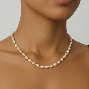 Può includere: Una delicata collana di perle con perle ovali e piccole perline dorate. La collana è indossata intorno al collo, mettendo in risalto la lucentezza naturale delle perle e il tono caldo dell'oro. Un gioiello classico.
