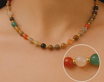May include: A beaded necklace featuring a variety of colorful gemstones. The necklace has a gold-colored clasp and small gold-colored beads between each gemstone. The gemstones are in shades of red, orange, yellow, green, brown, and black.