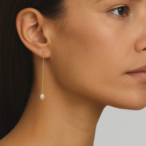 Può includere: Un singolo orecchino a catena d'oro con una piccola perla bianca all'estremità. L'orecchino pende dall'orecchio di una persona. Lo sfondo è di colore neutro.
