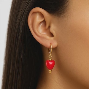 Può includere: Orecchini a cerchio color oro con un ciondolo a forma di cuore rosso. Gli orecchini hanno una piccola perla dorata nella parte inferiore del cuore e una perla dorata nella parte superiore del cuore. Gli orecchini sono indossati sull'orecchio.