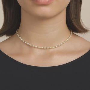 Può includere: Una delicata collana con piccole perle bianche crema intervallate da perline color oro. La collana è indossata intorno al collo, appena sotto la clavicola. Lo sfondo è di un tono neutro, evidenziando il gioiello.
