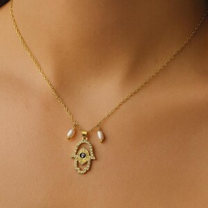 May include: Gold-toned necklace featuring a Hamsa hand pendant adorned with clear crystals and a central eye design. Two small, white pearl drops dangle from the pendant. The delicate chain is visible against the skin.