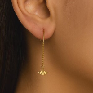 May include: A gold-colored earring with a chain and an eye-shaped pendant. The earring is worn in the ear and hangs down. The pendant has a stylized eye design with a sunburst pattern.