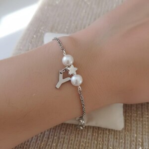 Può includere: Bracciale color argento con una catena sottile, due perle finte, un ciondolo a forma di stella e un pendente a forma di lettera "Y". Il bracciale è indossato su un polso, mettendo in risalto il suo design elegante e la sua sottile lucentezza.