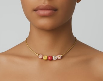 May include: A gold-toned necklace with the word "QUEEN" spelled out in colorful, round beads. The beads are pink, yellow, red, and white. The necklace is worn around the neck.