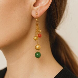Può includere: Orecchini pendenti color oro con una serie di perline sferiche colorate. Gli orecchini hanno un delicato design a catena, con perline nelle tonalità dell'oro, del rosso, del giallo e del verde. Gli orecchini sono progettati per essere indossati con orecchie forate.