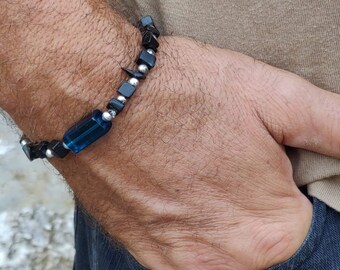 Puede incluir: Una pulsera de cuentas con una gema azul rectangular, cuentas cuadradas negras y detalles plateados. La pulsera se muestra en una muñeca, mostrando su diseño y contraste de color. Una joya elegante.