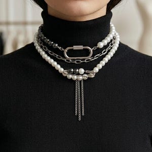 Può includere: Una collana a strati con perle bianche, catene in metallo argentato, una grande chiusura a moschettone rettangolare e un ciondolo a forma di spilla da balia con tre sottili catenelle argentate pendenti, indossata sopra un maglione nero a collo alto.
