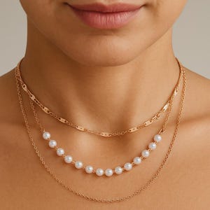 May include: A layered gold-toned necklace with three strands. The top strand has a chain-link design, the middle strand features a row of white pearls, and the bottom strand is a delicate chain. The necklace is worn around the neck.
