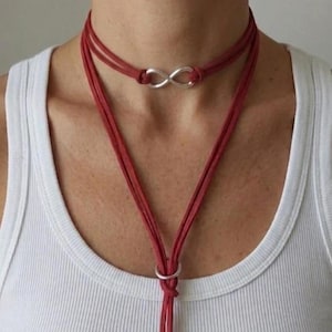 Può includere: Una collana girocollo in camoscio rosso con un ciondolo a simbolo infinito argentato. La collana presenta un design a strati con un lungo filo pendente e un dettaglio ad anello argentato. La collana è indossata sopra una canotta bianca.