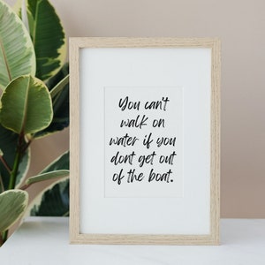Op de afbeelding: Een ingelijst print met een witte achtergrond en zwarte tekst die "You can't walk on water if you don't get out of the boat." luidt.