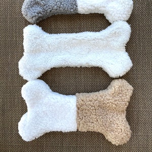 Puede incluir: Tres juguetes de peluche para perros con forma de hueso. El hueso superior es gris y blanco, el hueso del medio es blanco y el hueso inferior es blanco y marrón.