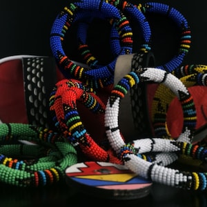 May include: A collection of colorful beaded bracelets in green, red, white, blue, and yellow. The bracelets are made of small beads and are arranged in a circle.