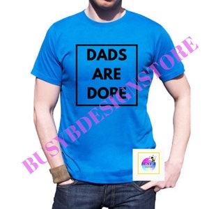 May include: A blue t-shirt with the words "DADS ARE DOPE" in a black square. The shirt is being worn by a person. The image also includes a small logo in the bottom right corner.