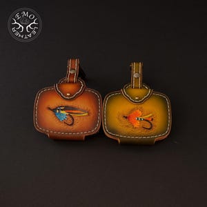 May include: Two leather fly fishing reel cases. One is brown with a blue and yellow fly design, the other is olive green with an orange fly design. Both have a top flap with a snap closure and white stitching.